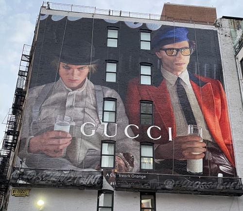 A huge mural on the exterior of a city building shows two strange-looking young men holding glasses of milk and wearing hats. G U C C I is prominent across the lowest third of the mural.